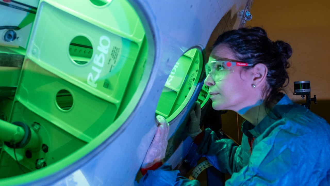 A woman wearing safety glasses and a blue lab coat peers into a circular portal of a large piece of industrial or aerospace machinery. The interior of the machine is bathed in a vibrant green light, highlighting a structural frame with circular cutouts.