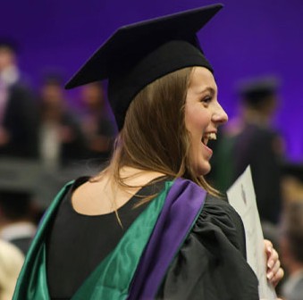 A female student wearing a graduation gown and cap, looking excitedly off-camera as she holds up her degree certificate