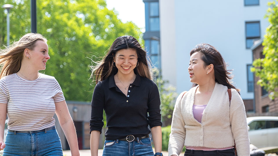 Three students walking outside with halls of residence in the background.