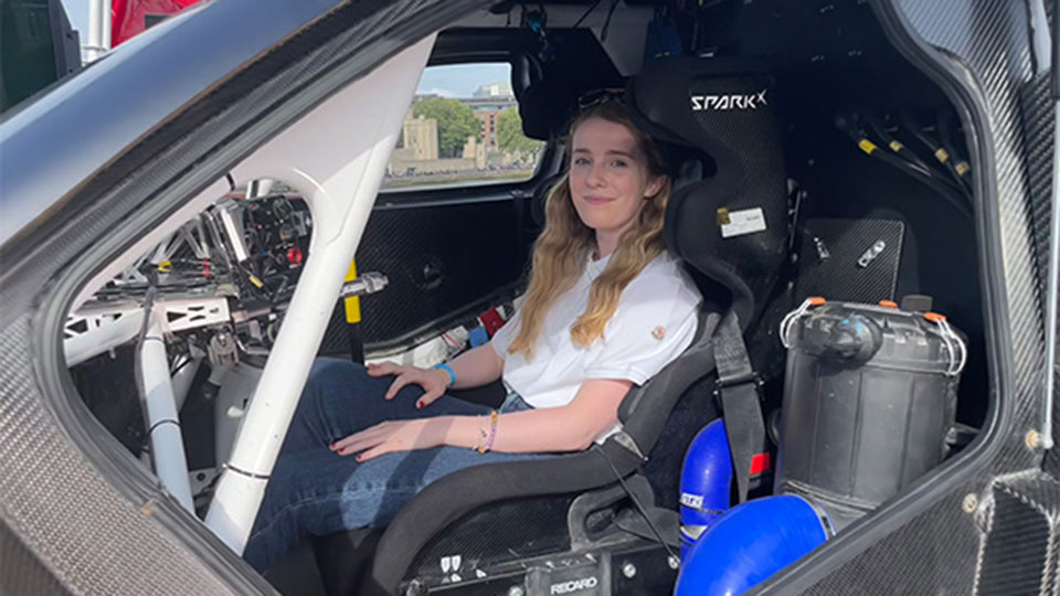 A woman with long blonde hair sits in the racing seat of a high-performance vehicle's cockpit. The interior features carbon fiber paneling, a Spark racing seat, and visible mechanical components. She is wearing a white t-shirt and jeans, looking toward the camera with a slight smile.