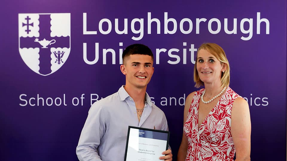 Student Fin holding an award certificate from Jan Godsell, Dean of the School of Business and Economics. There is a large purple banner behind them displaying the Loughborough University logo.