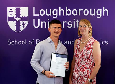 Fin Dearsly receiving the School of Business and Economics Dean's Award for Enterprise 2022. He is holding the award and standing beside the Dean, Jan Godsell. There is a large University-branded purple banner behind them.