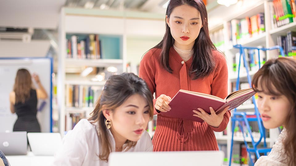 Three students looking at a laptop screen with shelves of books in the background