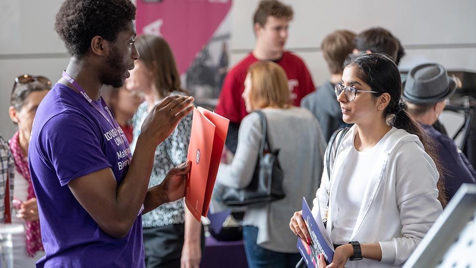 A prospective student talking to a student ambassador who is holding a brochure and wearing a purple t-shirt. More open day guests are in the background.