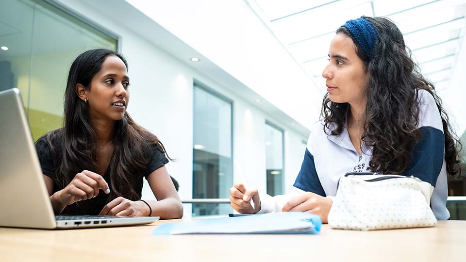 Two students in conversation in a bright campus study space with glass-fronted rooms and a skylight overhead
