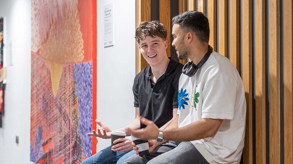 Two students sitting and talking in a modern campus building, with artwork and wooden wall panelling in the background.