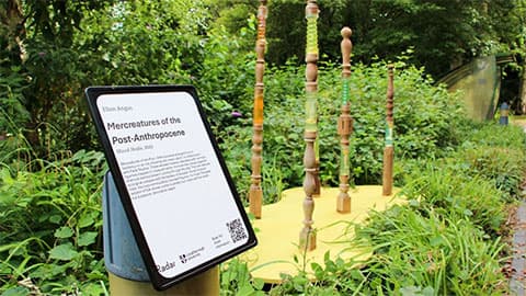 Outdoor sculpture with carved wooden pillars and green glass inserts, next to an informative sign titled 