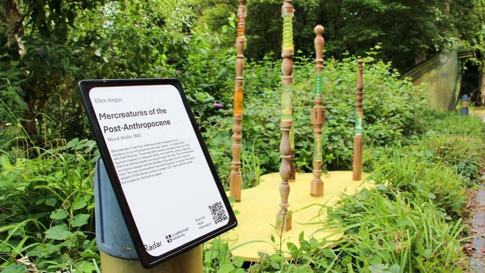 Outdoor sculpture with carved wooden pillars and green glass inserts, next to an informative sign titled 
