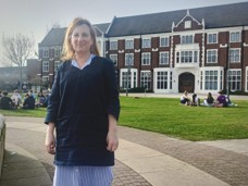 A woman stands smiling in front of a campus building. Students sit on grass, enjoying a sunny day.