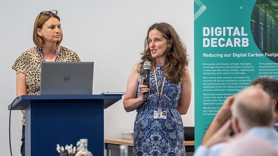 Two women are standing near a podium with a laptop. One speaks into a microphone. A 