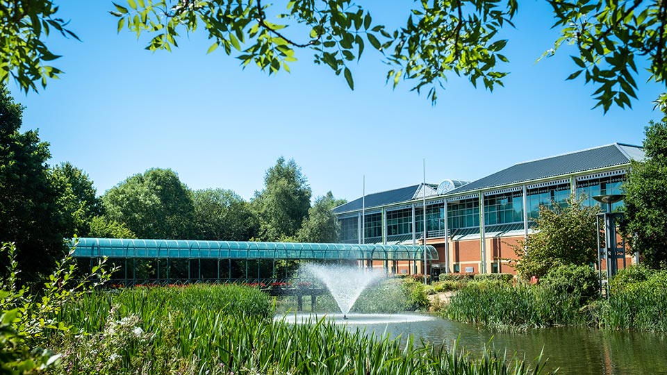 A modern building with large windows and a blue roof stands next to a serene pond with a fountain.