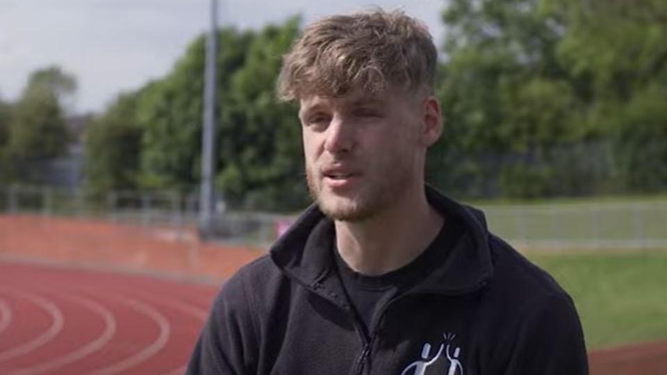 Photo of current student Joe, founder of theathlete place, speaking to the camera in front of the outside running track at the Paula Radcliffe Stadium on campus