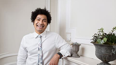 Jason is standing against a mantle piece smiling against a plain background with a plant pot in the bottom right hand corner.