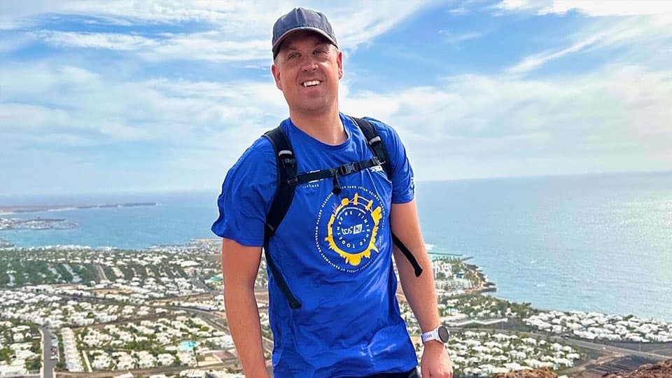 Paul is standing on a high mountain and you can see a village of houses in the background. He is wearing walking gear and he is smiling at the camera.