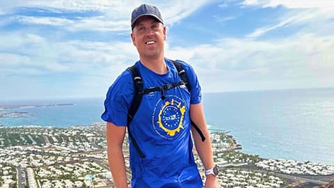 Paul is standing on a high mountain and you can see a village of houses in the background. He is wearing walking gear and he is smiling at the camera.