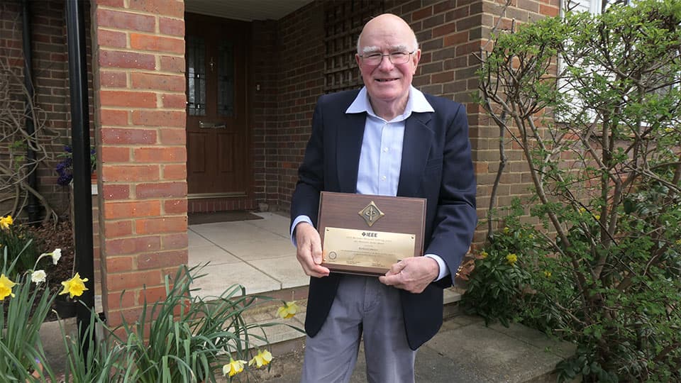 Richard is wearing a suit and is standing outside smiling holding his award.