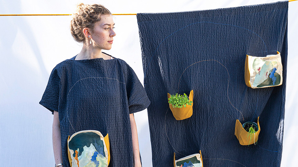 Photo of Alice Burnhope wearing 'Nature to go' whilst stood next to wall hanging, 'Hang Out'.