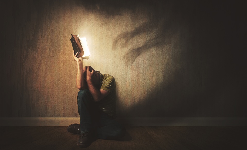 Man sat on the floor hiding his face in a book as a shadow-like ghost creeps across the wall behind him. 