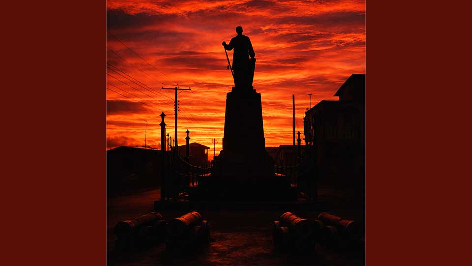 A silhouette figure stood on top of a pillar in an urban setting during sunset