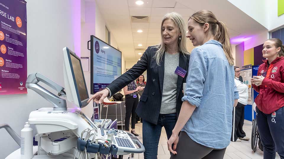 Two women stand together in front of a computer, engaging in conversation and pointing at the screen.