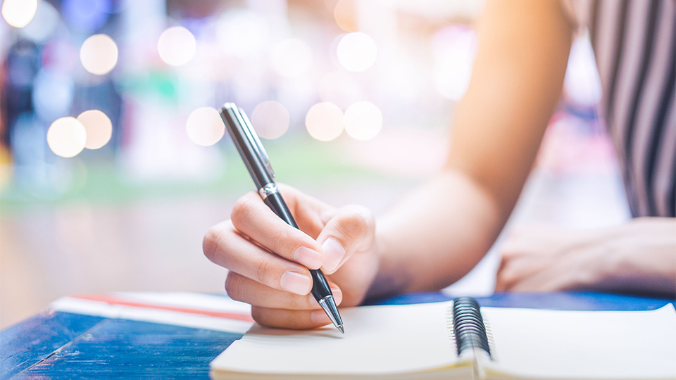 photo of somebody holding a pen and writing in a notebook on a table