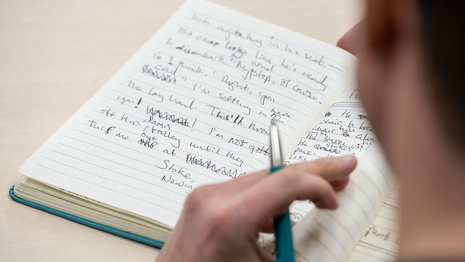 photo of somebody holding a pen and writing in a notebook on a table