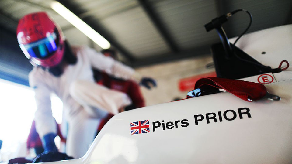 Close up image of Piers' racing car with his name and the United Kingdom flag printed on. He is in the background with racing gear and helmet on