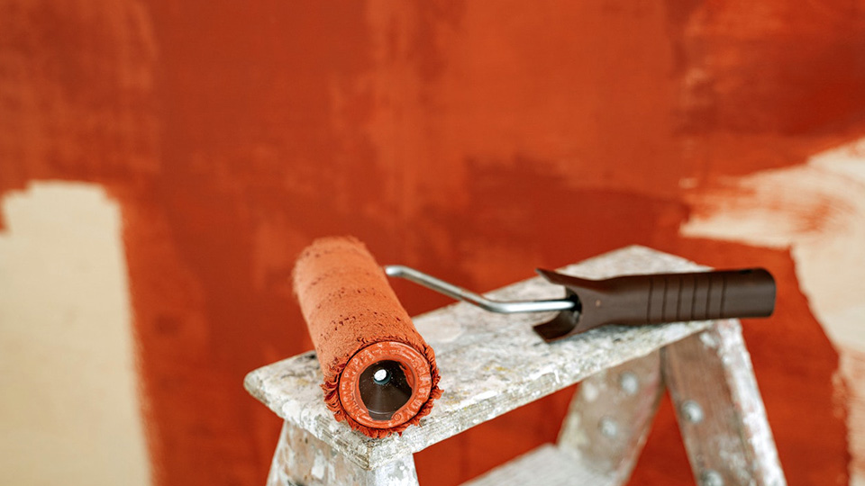 A photo of a paint roller lay on a bench with red paint on, and a red wall behind it