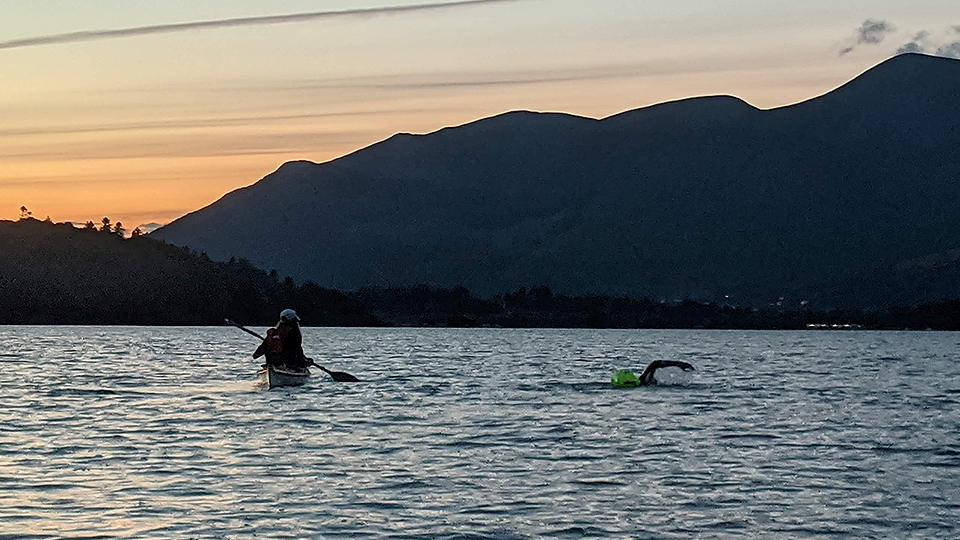 Photo of Lboro academic Danny Longman swimming across a lake in the Lake District, with a mountain backdrop behind him. Image also includes one of his support team on a kayak 