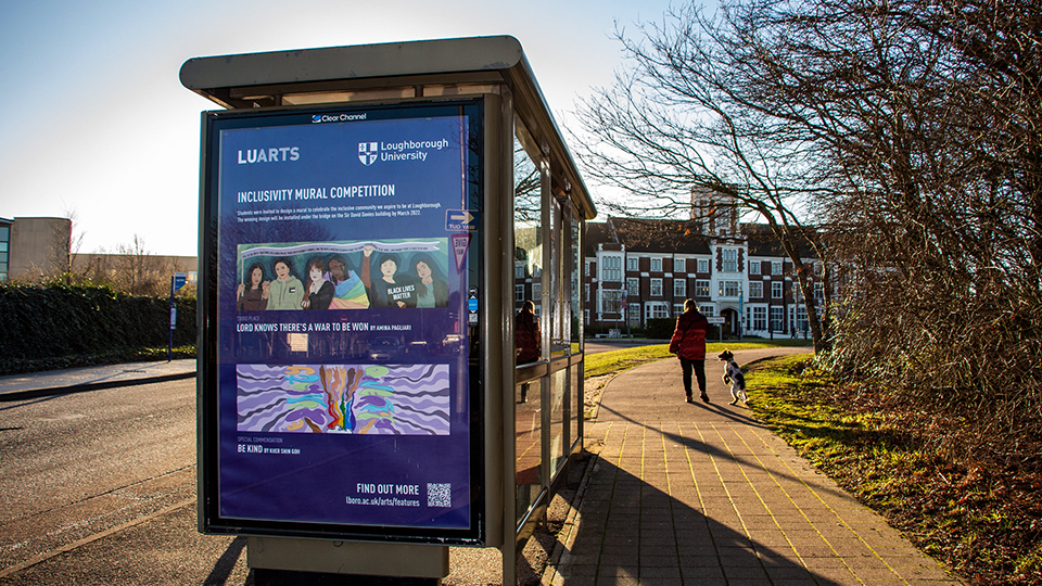 Photo of competition winner designs being shown in a bus shelter poster on campus, with Hazlerigg Building in the background and a person with a dog walking past