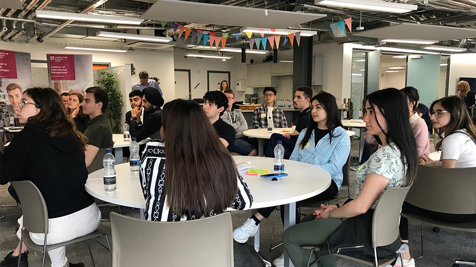 Photo of students sat on tables in a London campus room listening to someone talking. The room is decorated with colourful bunting along the ceiling
