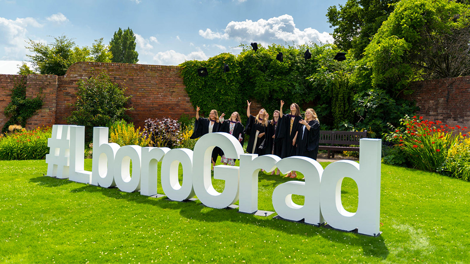 Photo of group of women graduates standing in front of big white standing letters that say #LboroGrad on the lawn on campus