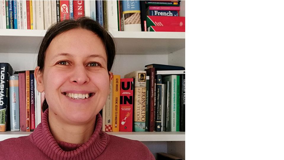 Headshot of Kinga with her hair tied up wearing a pink roll neck jumper, smiling at the camera with a bookshelf behind her