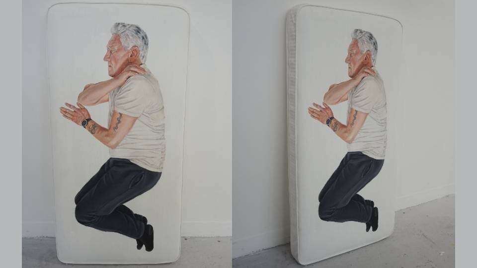 A small white cot mattress leaning against a white wall, painted on it is an older person lying on their side with their eyes closed.
