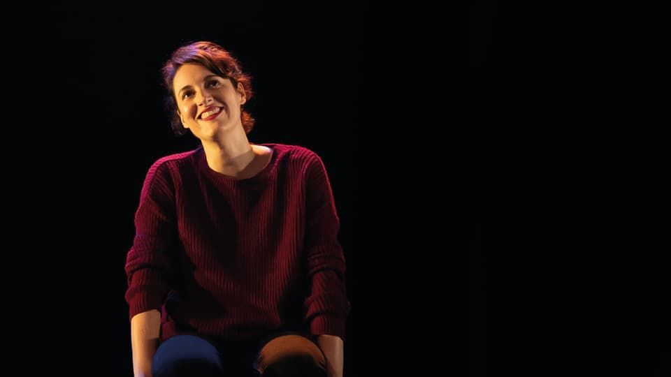 Phoebe Waller-Bridge in a spotlight, sitting on a chair looking upwards and smiling on a dark stage.