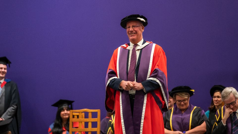 A man standing and smiling at a graduation ceremony wearing an academic gown and hat.