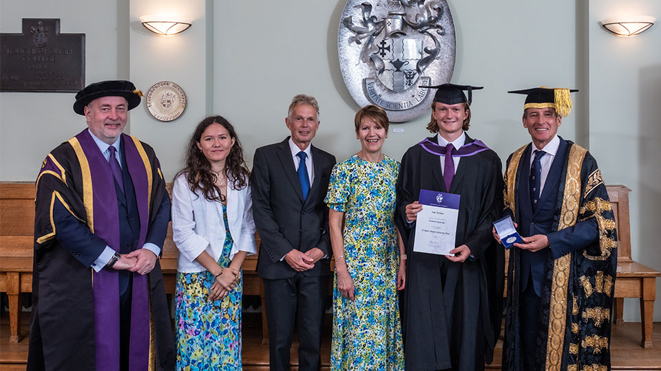 Four men and two women standing in a line. The two men on the right are holding a medal and a certificate. Three men are in academic robes. The other man and two women are wearing formalwear.