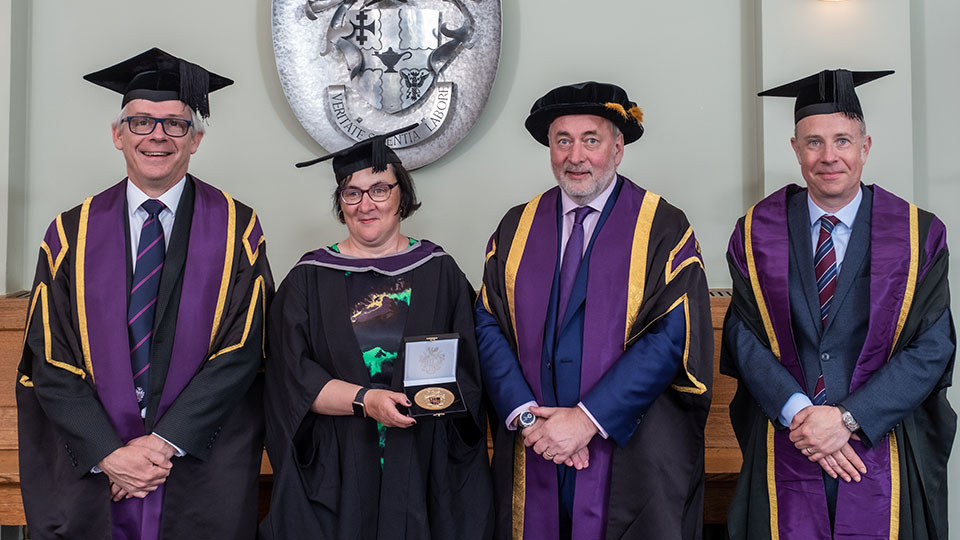 Three men and one woman in academic robes. The woman is holding a medal 