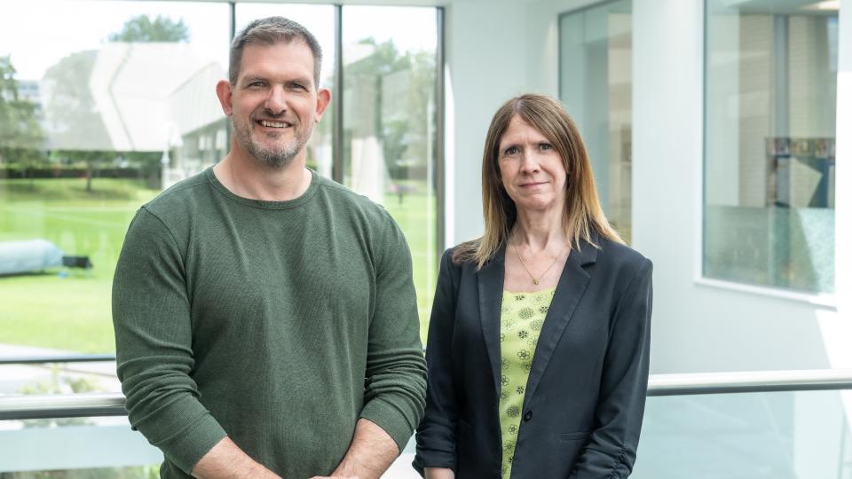 Photo of Dr Ash Casey and Professor Lorraine Cale stood next to each other looking at the camera, in front of a window. Ash is wearing a green jumper and Lorraine is wearing a yellow patterned top with a black blazer. 