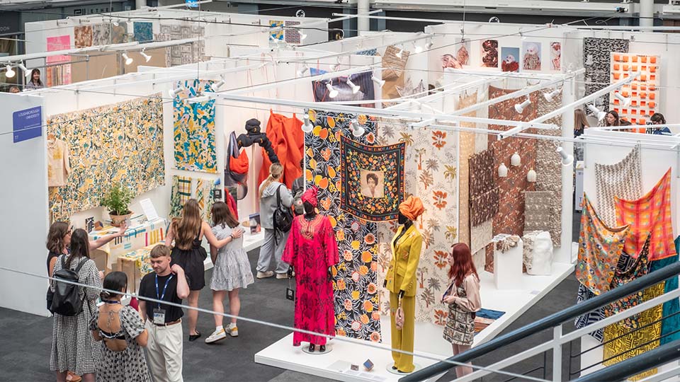 A photo taken high up of the Loughborough stands at Week 1 of New Designers. You can see students and attendees talking, as well as mannequins wearing garments and large pieces of fabric on display which has been created by the students