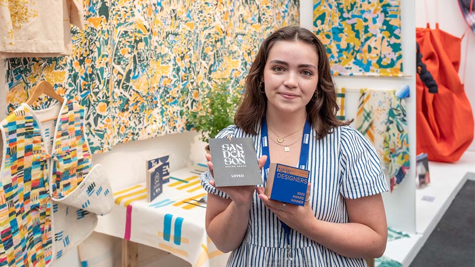 A photo of student Catherine Owen-Milsom, holding the Sanderson Design Award trophy, smiling and looking at the camera