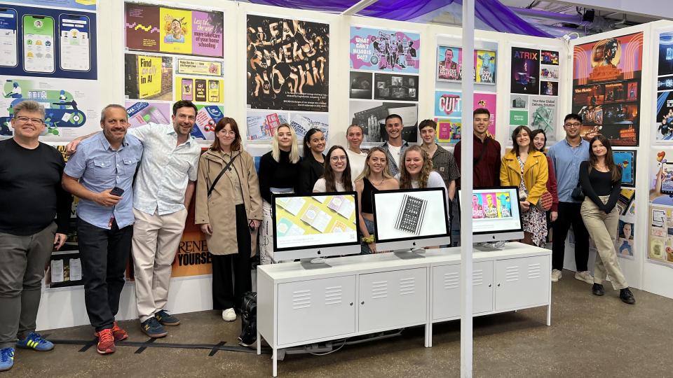 Group of Loughborough staff and graduates standing in front of the D&AD New Blood exhibition which consisted of their graphics and illustration work on gallery walls and across three computer screens. 