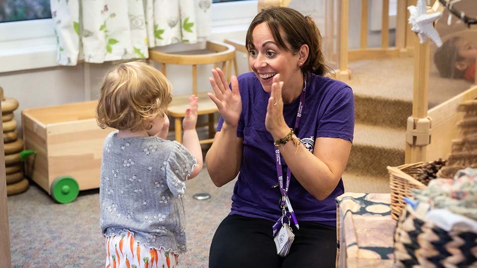 Photo of a nursery worker wearing a purple top and lanyard, smiling and clapping hands with a toddler at the nursery. Various toys and furniture surround them in a room