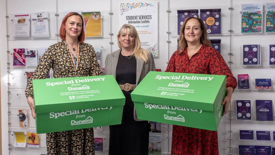Three people standing in front of a wall of Student Services information leaflets. Two of them are holding big green boxes that have 'Special Delivery from Dunelm' written on top.