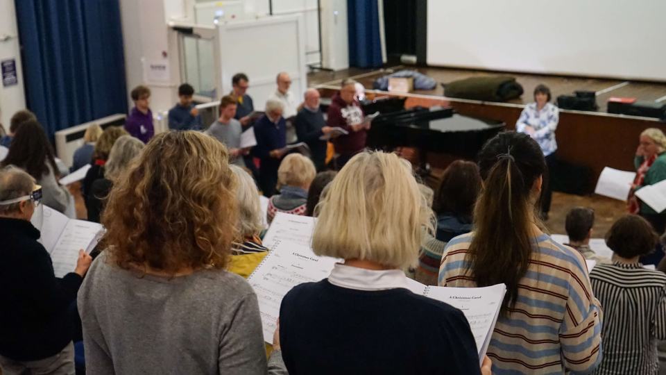 At a University Choir rehearsal, a group of people standing together reading lyrics from music books.