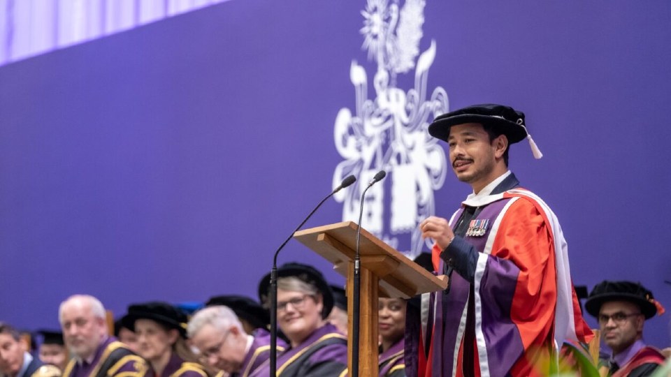 Nims Purja speaking on stage, standing behind a lectern, wearing graduation wear..