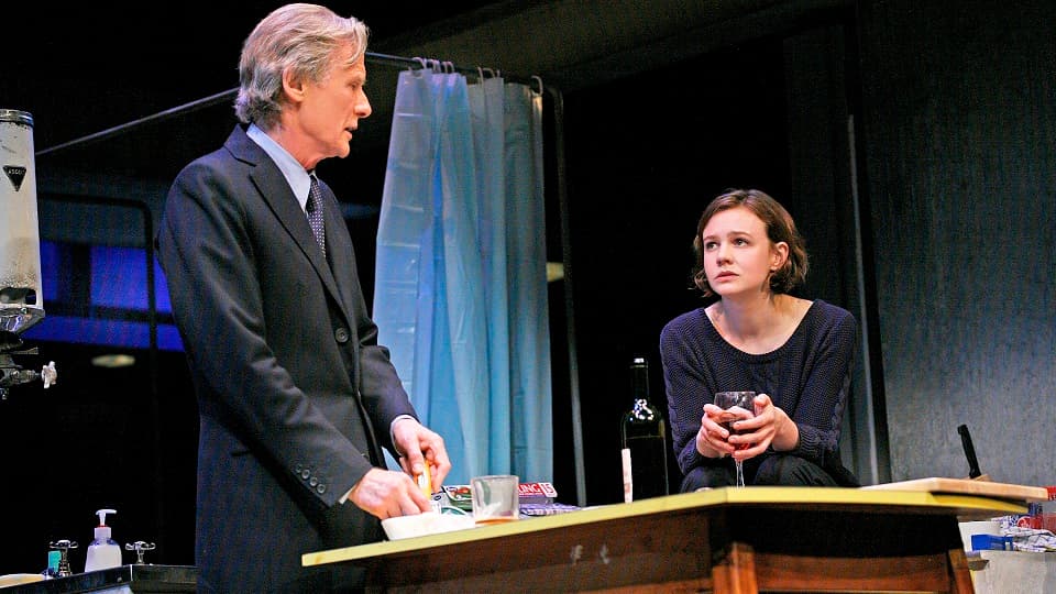 Two people talking at a table on a theatre set, one is standing wearing a suit, the other is sitting with their hands round a glass of red wine. 