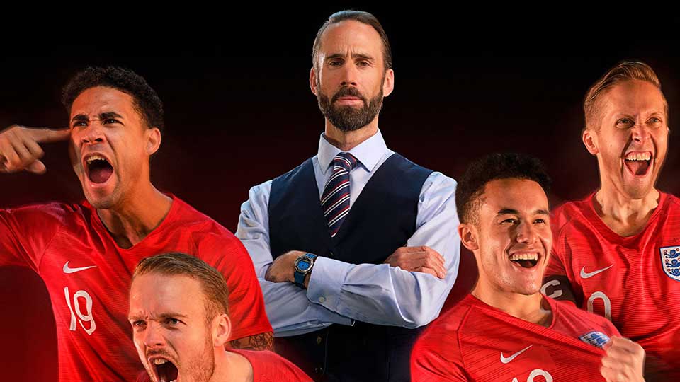 A person wearing a shirt and tie is standing with their arms folded between four football players wearing red England football shirts and celebrating.