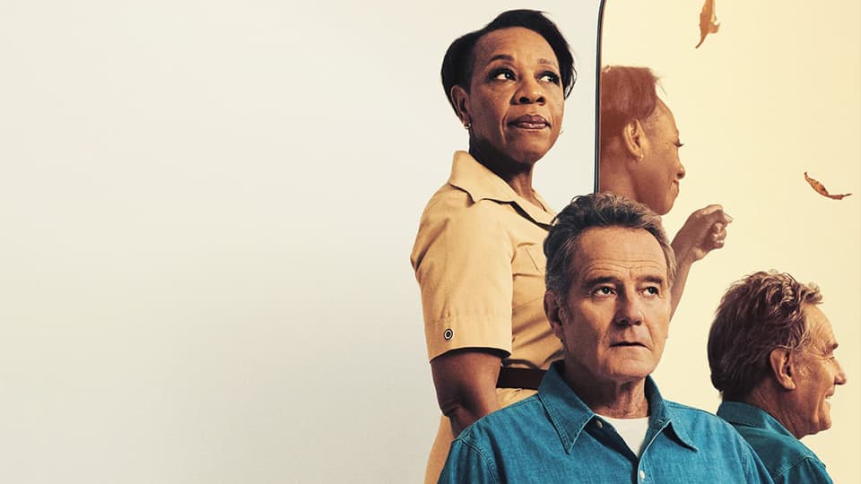 A married couple, one person stood behind one person sat, both in front of a mirror looking left