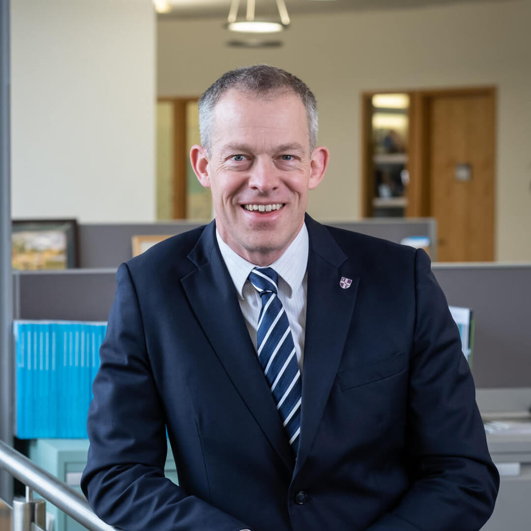 Male Dean of School smiling at the camera while standing next to a staircase in a well-lit room, wearing a suit.